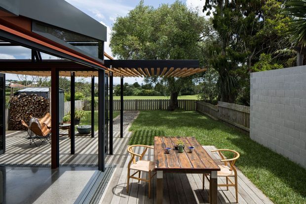 Contemporary residential outdoor dining area beside a glazed living space, with a timber deck, rustic timber dining table and chairs, and a slatted timber pergola. Black-framed floor-to-ceiling glazing opens onto a landscaped backyard with lawn, fencing, mature trees, and a light concrete-block boundary wall. The house has a strong modern geometric form with dark cladding and warm timber accents.