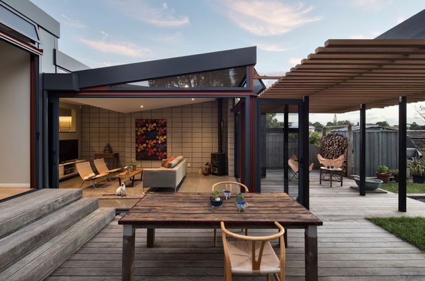 A contemporary outdoor entertaining area with a timber deck, dining table and chairs in the foreground, and a covered pergola to the right. The space opens onto a glazed living room featuring a low sofa, lounge chairs, tiled walls and a fireplace or media wall. Dark metal framing contrasts with warm timber decking and pergola slats, creating a seamless indoor-outdoor connection.