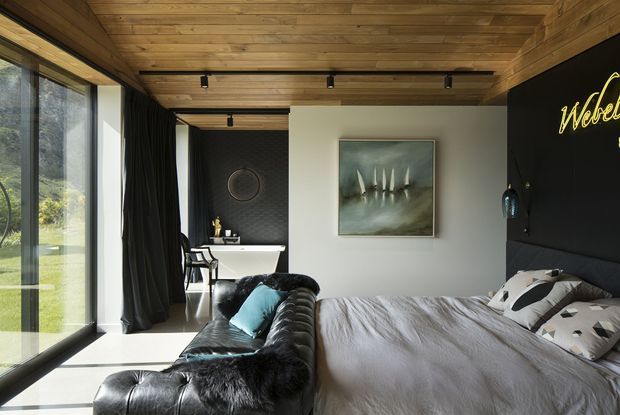 A stylish bedroom with a large bed, dark leather sofa, and floor-to-ceiling glazing opening toward a garden. The room features a warm timber-lined ceiling, light walls, dark curtains, black feature wall, wall-mounted artwork, and integrated track lighting. A compact desk or vanity area is visible at the rear.