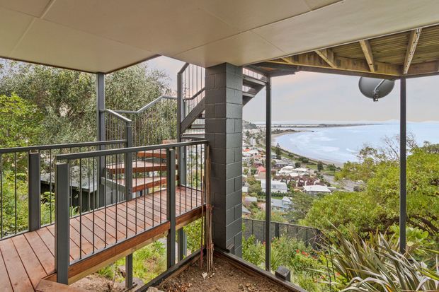 Elevated outdoor deck and covered balcony overlooking a coastal bay and hillside residential neighbourhood. The space features timber decking, dark metal balustrades and posts, a concrete block structural column, and a partially exposed timber roof structure, with stairs connecting to a lower level.