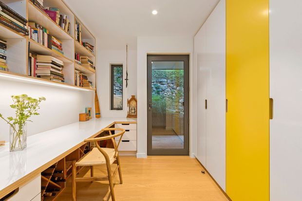 Bright compact home office or study with a long white built-in desk, open timber shelving filled with books, woven wooden chairs, integrated under-shelf lighting, pale timber flooring, and a striking yellow built-in cupboard. A glazed door opens to a small exterior space with stone landscaping visible beyond.