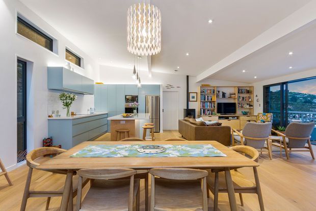 Open-plan contemporary kitchen, dining and living area with pale blue-grey cabinetry, a central island, extensive light timber flooring, and a large timber dining table in the foreground. The living zone includes built-in timber shelving, a television, upholstered seating and large glazed doors opening toward a scenic outdoor view. A sculptural pendant light hangs above the dining area, with recessed ceiling lighting throughout.