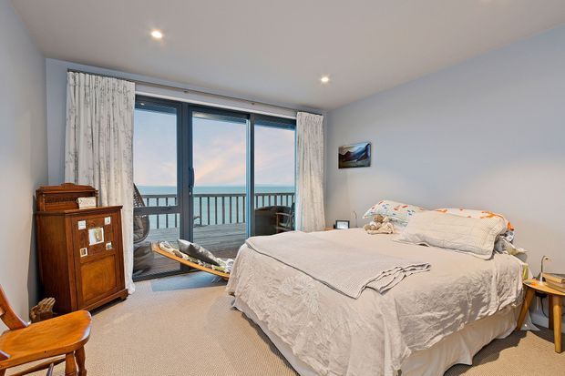 A bright bedroom with a large bed dressed in light neutral linens, pale walls, carpeted flooring, and full-height glazed doors opening to a balcony with a railing and ocean or sky views. White curtains, recessed ceiling lights, a timber cabinet, and a small side table complete the room.