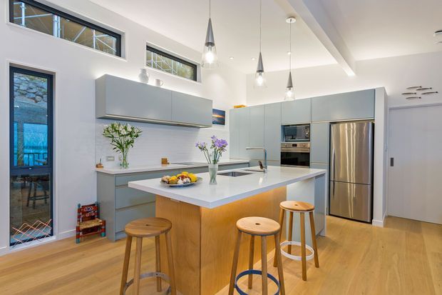 Bright contemporary kitchen with a central timber-based island, white benchtop, pale blue-grey cabinetry, stainless steel appliances, timber flooring, and pendant lighting. High-level windows and a glazed door bring in natural light, while stools provide casual island seating.