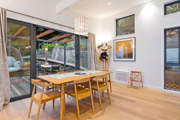 Bright contemporary dining area with a long light-wood dining table and matching chairs, set beside expansive dark-framed glazing and sliding doors opening to a timber deck and landscaped outdoor space. White walls, pale timber flooring, a sculptural pendant light, framed artwork, and a compact wall heater create a clean, warm, indoor-outdoor setting.