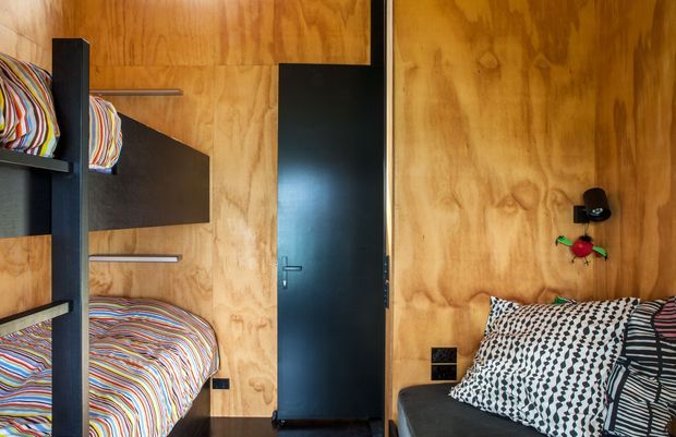 Compact bedroom with warm plywood wall and ceiling surfaces, built-in bunk beds on the left, a lower bed in the foreground, and dark charcoal built-in shelving, partitions, door, and furniture. Bold multicoloured striped and geometric bedding adds a playful graphic character.