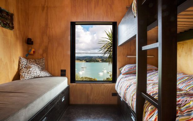 Compact timber-lined bedroom with two built-in sleeping areas, including a lower bed at left and stacked bunk-style beds at right. Dark built-in joinery and carpet contrast with the warm wood walls. A central black-framed window overlooks a calm coastal inlet with boats and hills, while colourful striped bedding adds visual interest.
