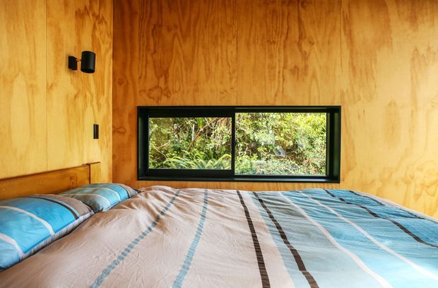 Compact bedroom with warm plywood wall and ceiling surfaces, a low horizontal black-framed window overlooking dense greenery, and a bed filling most of the room. The bed has blue, grey, white, and brown striped bedding, while a small black wall light is mounted beside the window.