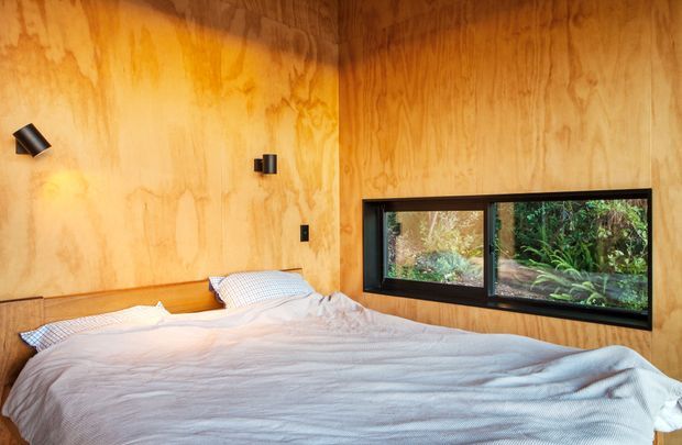 A compact bedroom clad in warm natural timber wall panelling, with a low built-in timber bed surround, pale bedding, and a horizontal black-framed window overlooking dense greenery. Small black wall-mounted reading lights and switches provide simple functional detailing. The room has a warm, cozy cabin-like atmosphere and appears to have a low, standard-height ceiling.
