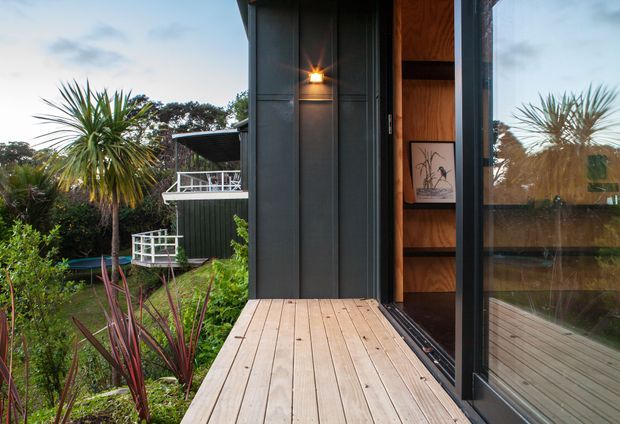 Exterior view of a compact modern dwelling entrance, showing a timber deck leading to a tall glazed door and dark vertical cladding. The open doorway reveals warm timber shelving and a framed artwork inside. Lush landscaped planting surrounds the deck, with another elevated deck or balcony visible in the background.