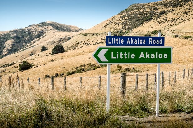 A roadside directional sign for Little Akaloa Road and Little Akaloa stands beside a wire fence in a dry, rolling mountain landscape under a clear blue sky.