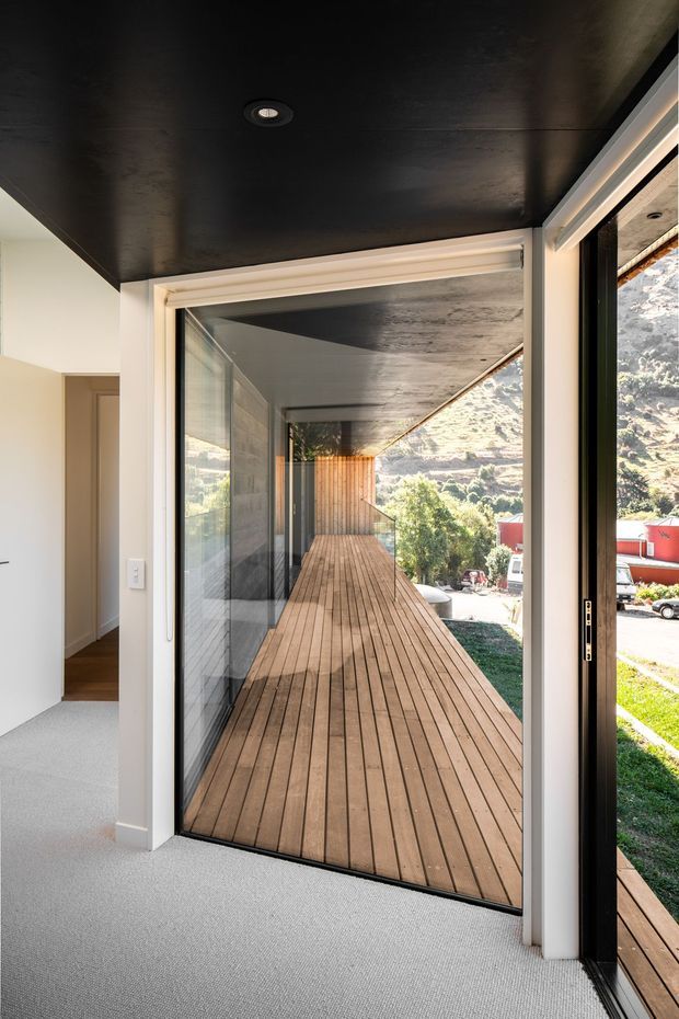 Interior view looking through large glazed sliding doors onto a narrow timber deck or balcony. The space has light carpet, white walls and trim, a dark painted ceiling with recessed lighting, and black-framed glazing. The deck extends along the exterior toward a timber-clad wall, with a sunlit mountainous or hilly landscape visible beyond.