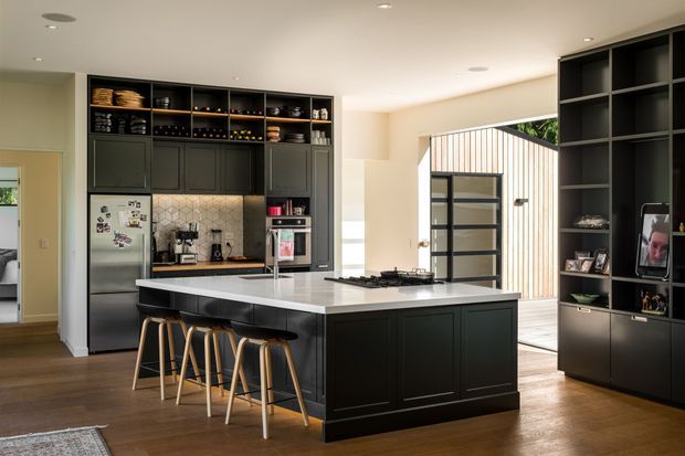 Contemporary open-plan kitchen with a large black island, integrated seating, dark full-height cabinetry, open display shelving, stainless-steel appliances, and a pale stone benchtop. Warm timber flooring contrasts with the dark joinery, while a glazed opening provides views toward a light-clad outdoor area. The space is clean, refined, and moderately bright.