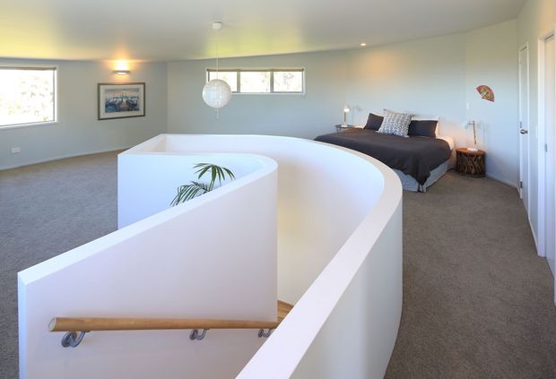 A spacious contemporary bedroom with a distinctive curved white stairwell or mezzanine balustrade in the foreground. The room has grey carpet, pale grey walls, a low horizontal window, a double bed with dark bedding, simple bedside furniture, recessed lighting, and a spherical pendant light. A timber handrail follows the curved stair opening, with a small plant visible below.