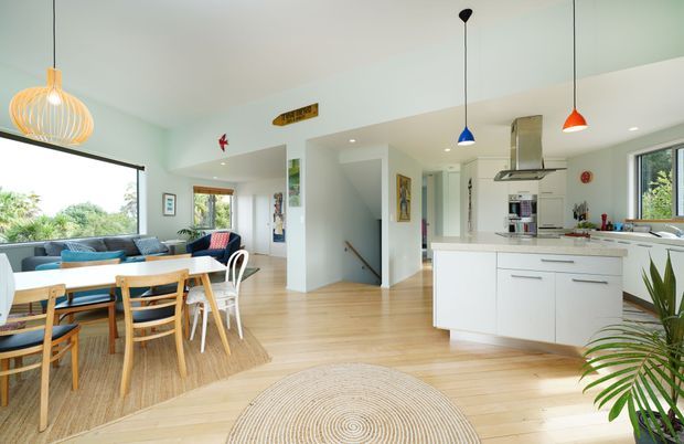 Bright open-plan residential interior combining a kitchen, dining area, and living room. The space features white cabinetry, light timber flooring, expansive glazing, colourful pendant lights, a timber dining table with mixed seating, and a compact lounge area overlooking greenery. The overall feel is airy, casual, and contemporary with warm natural materials.