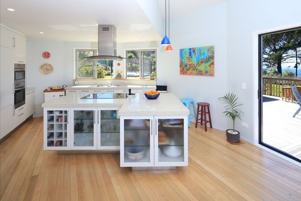 Bright, open-plan contemporary kitchen with white built-in cabinetry, a large central island with glass-front storage, integrated appliances, timber flooring, pendant lighting, and a stainless-steel rangehood. Large windows and a glazed door open to a timber deck, while colourful artwork and stools add accents to the light-filled space.