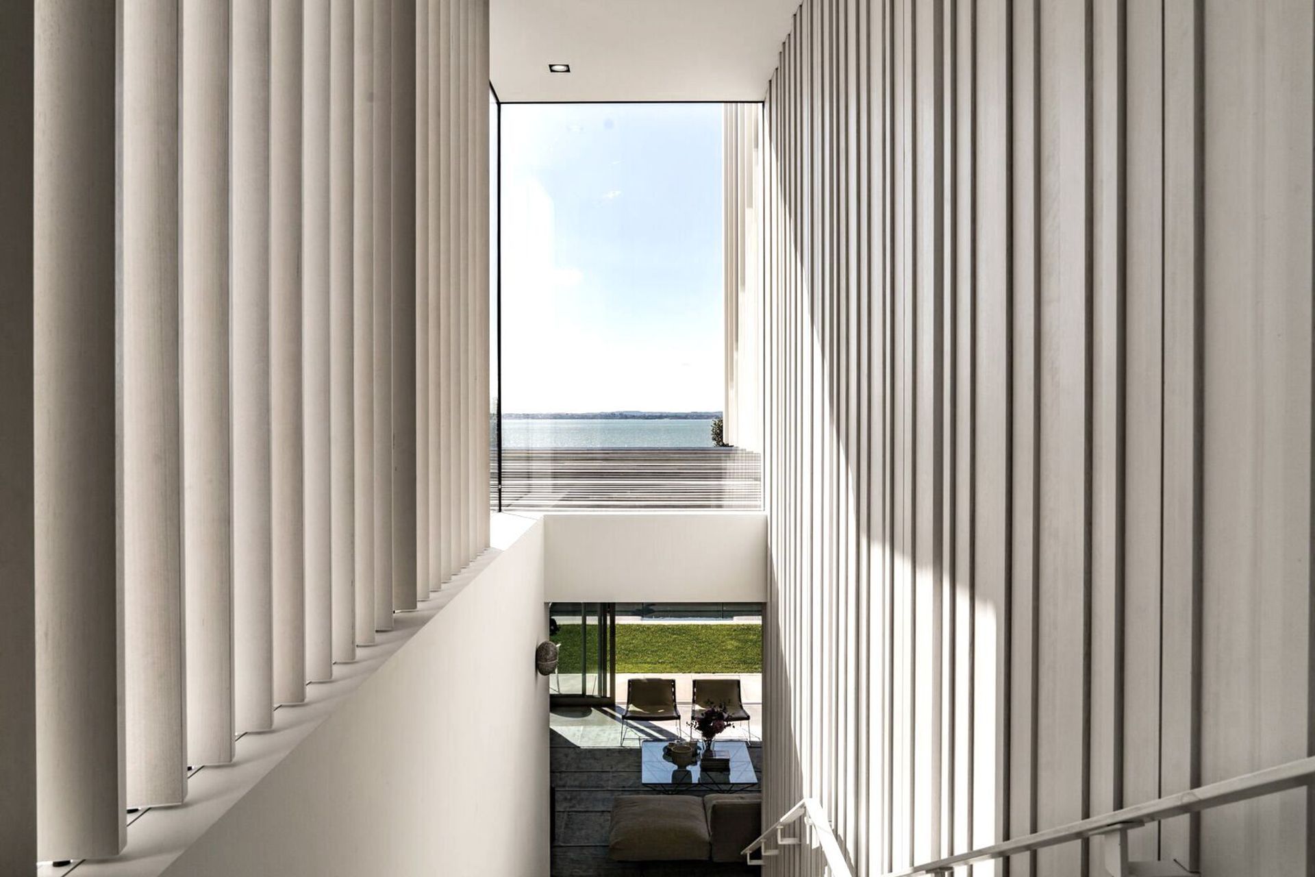 The main central staircase draws the eye to a feature window above, which frames a view of the sea, and then it opens up into the living area, below, and the landscaped pool area.