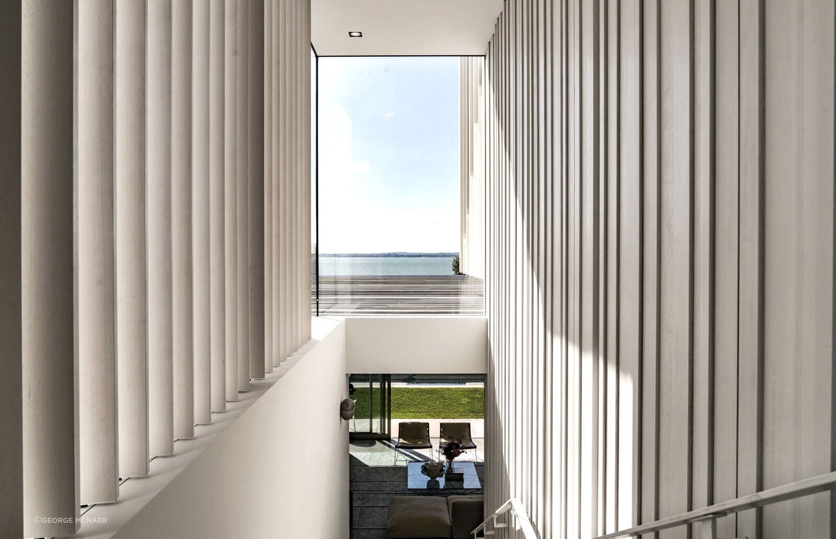 The main central staircase draws the eye to a feature window above, which frames a view of the sea, and then it opens up into the living area, below, and the landscaped pool area.