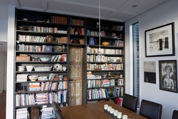 A dining room dominated by a full-height dark built-in library wall filled with books, ceramics, framed objects, and a central distressed timber cabinet door. A large timber dining table with dark upholstered chairs occupies the foreground, while framed artwork hangs on the light wall beside a tall window. The room has a refined, collected, book-lined character with decorative coffered ceiling detailing.