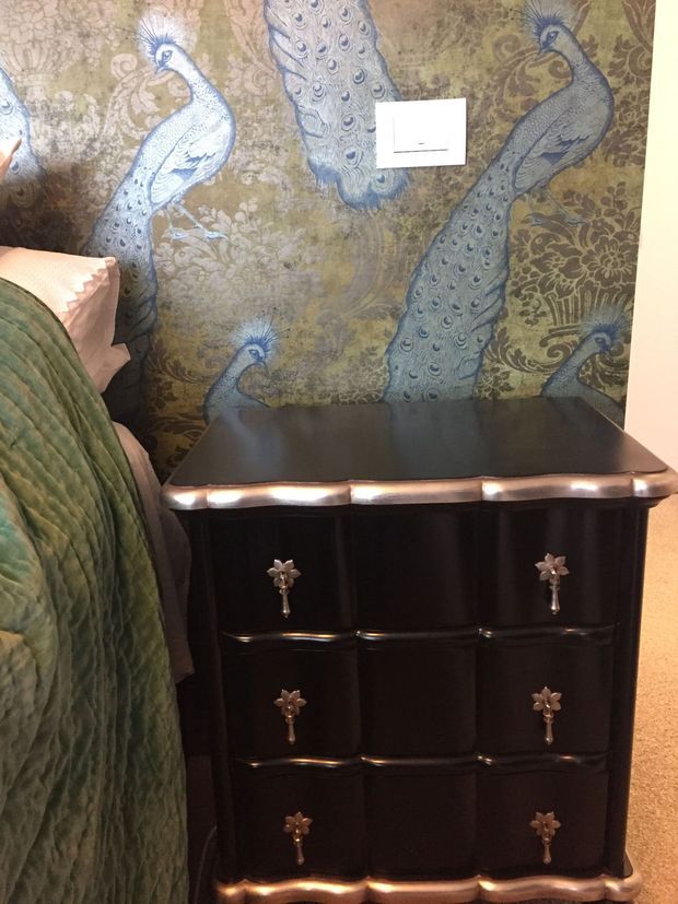 Close-up interior view of a dark, glossy chest of drawers beside a bed, with ornate metallic drawer pulls and curved trim. The wall behind features a dramatic peacock-patterned decorative wallpaper in muted green, brown, and blue tones. A quilted green bedspread and pale pink bedding are partially visible at the left.