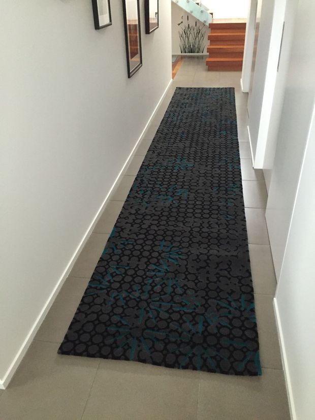 A narrow, bright residential hallway with white walls and pale tiled flooring, featuring a long dark patterned runner rug with teal accents. Framed artwork lines the left wall, while timber floating stairs and a decorative feature wall are visible at the far end.