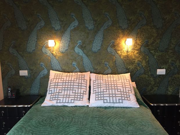 A compact bedroom viewed straight on, featuring a double bed with a green quilt and two patterned pillows. The bed is set against a dramatic dark green feature wall covered in a repeating peacock-pattern mural or wallpaper, flanked by two warm wall lights. Black built-in bedside cabinetry runs along both sides, creating a moody, richly patterned interior.