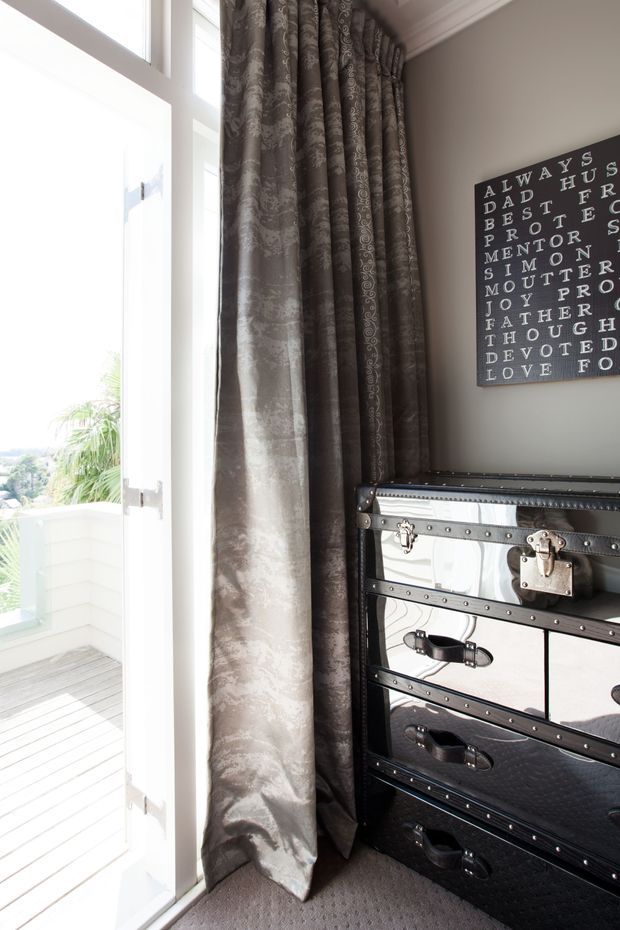 A bright interior corner beside a tall white-framed glazed door opening onto a small balcony. Floor-to-ceiling dark brown patterned curtains frame the opening, while a black mirrored trunk-style chest of drawers sits against a warm grey wall beneath a typographic wall plaque. The room has carpeted flooring and decorative ceiling moulding.