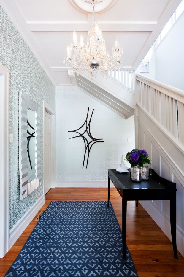 A bright, refined residential hallway and stair landing with pale patterned wallpaper, white decorative wall panelling and trim, a coffered ceiling, and a prominent crystal chandelier. A black console table with floral arrangements sits beneath the staircase, while a blue patterned runner contrasts with the warm timber floor. A decorative mirror and sculptural wall art add visual interest.