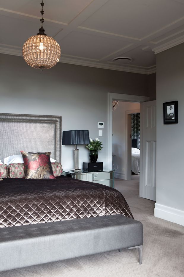 Elegant bedroom with a large upholstered bed, quilted dark brown bedspread, patterned accent cushions, mirrored bedside console, table lamp, and upholstered bench. The room features soft grey walls, plush carpet, ornate ceiling cornices, recessed ceiling detailing, and a decorative crystal globe pendant. An open doorway reveals an adjoining room or bathroom with patterned wall tiling.