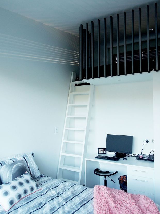 Compact, bright bedroom with a built-in loft sleeping platform accessed by a white ladder. The room includes a bed with patterned blue-grey bedding, a small built-in home office desk with computer and stool, white cabinetry, and a dark vertical-slat loft balustrade. The overall design is contemporary and space-efficient.