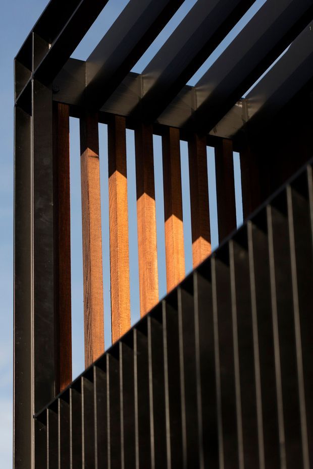 A dramatic architectural detail showing a dark metal stair or balustrade in the foreground, with vertical warm timber slats behind it and large black overhead roof beams creating a rhythmic pergola-like structure. Strong sunlight highlights the timber against a clear blue sky, producing deep shadows and geometric contrast.
