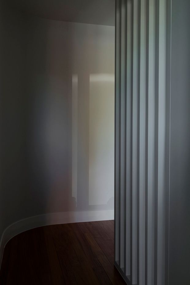 A dimly lit minimalist interior passage or room corner with dark timber flooring, smooth pale walls, and a prominent vertical fluted or slatted translucent partition on the right. Curved wall geometry and soft indirect light create a quiet, architectural atmosphere.