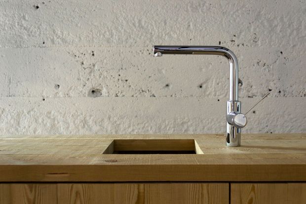 Close-up of a minimalist kitchen or utility-room sink integrated into a thick timber benchtop, with a rectangular undermount basin and a polished chrome gooseneck mixer tap. The wall behind is rough, textured off-white plaster or concrete, while vertical-grain timber cabinetry is visible below. The composition is architectural and material-focused, with no loose furnishings.