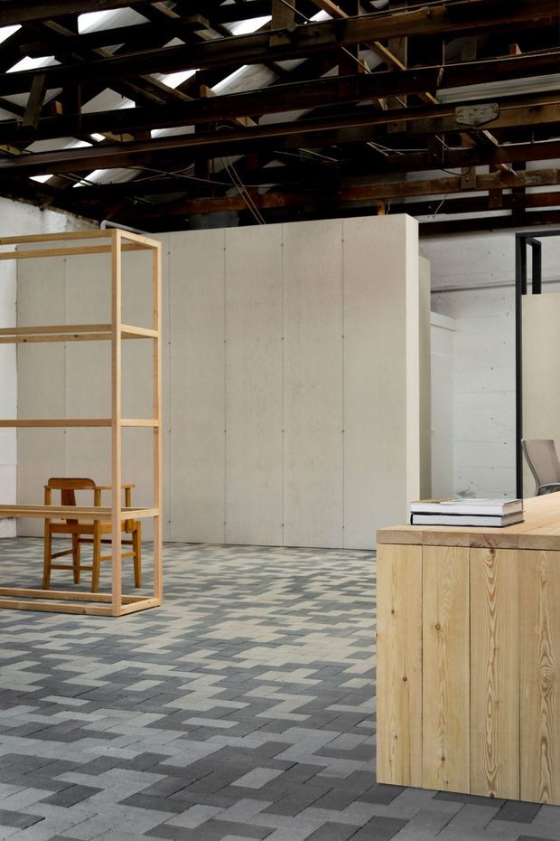 Interior of a converted industrial-style space with exposed dark timber roof structure and skylights, white paneled partitions, a freestanding pale timber shelving frame, a simple wooden chair and table, and a pale timber reception or storage counter. The floor is covered in a geometric pattern of rectangular grey tiles, creating a restrained contemporary workspace or showroom atmosphere.