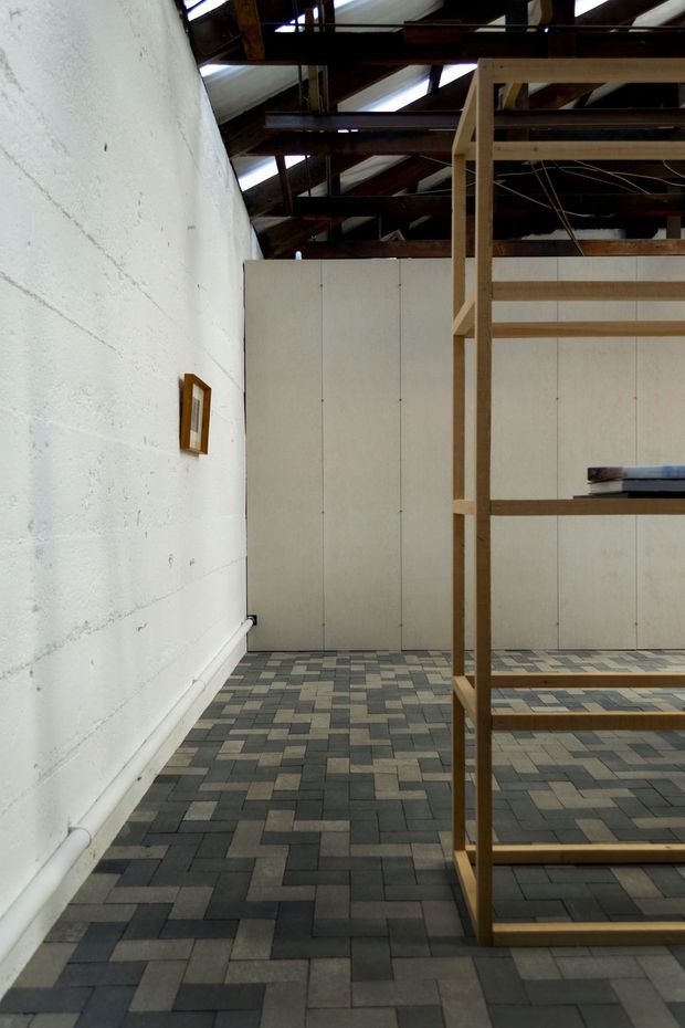 An austere, industrial interior with a white textured masonry wall, exposed dark timber roof structure and translucent roof panels. The space has a geometric grey, beige and charcoal tiled floor, pale vertical wall panels, and a tall open timber shelving or display structure in the foreground. The room appears sparsely furnished and utilitarian.