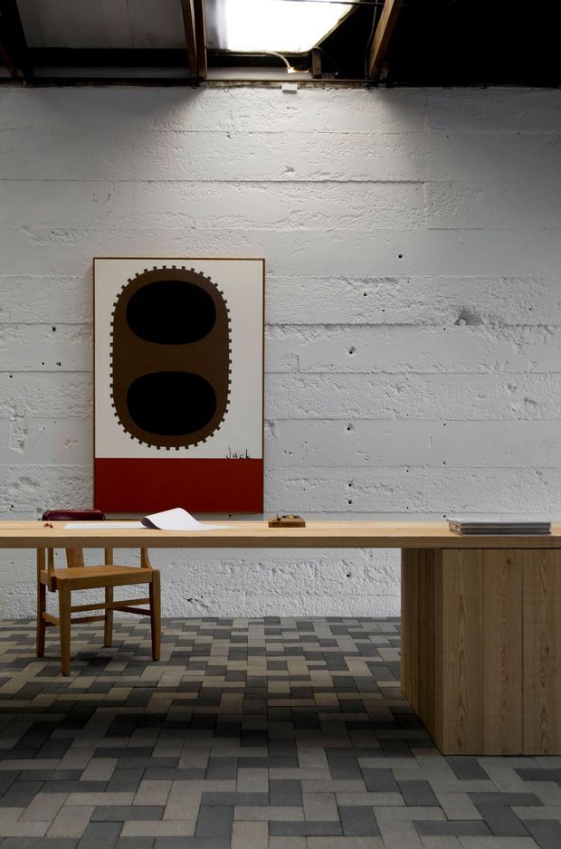 A moody, minimalist interior featuring a long light-wood desk or console with a timber chair, set against a rough white-painted masonry wall. A framed graphic artwork hangs above the desk, while the floor is finished in a grid of alternating grey and charcoal rectangular tiles. Exposed roof framing and a bright rooflight or skylight are visible overhead, contributing an industrial, gallery-like atmosphere.