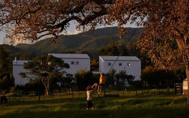 A rural residential scene at golden hour, viewed across a grassy yard toward two simple white rendered buildings with low-pitched metal roofs. Mature trees frame the view, with a forested mountain ridge in the background and two people playing in the foreground. The architecture is understated and contemporary, with small square windows and dark roof finishes.