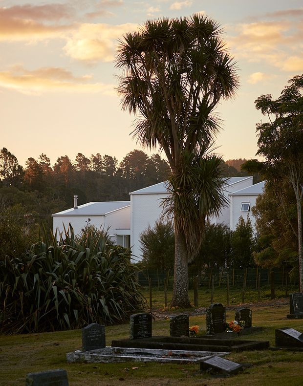 A landscaped outdoor scene at sunset, showing a tall cabbage tree in the foreground, dense native planting, and a modern white multi-level building with low-pitched roof forms in the background. Several dark memorial headstones and plaques occupy the foreground, suggesting a cemetery or memorial garden setting.