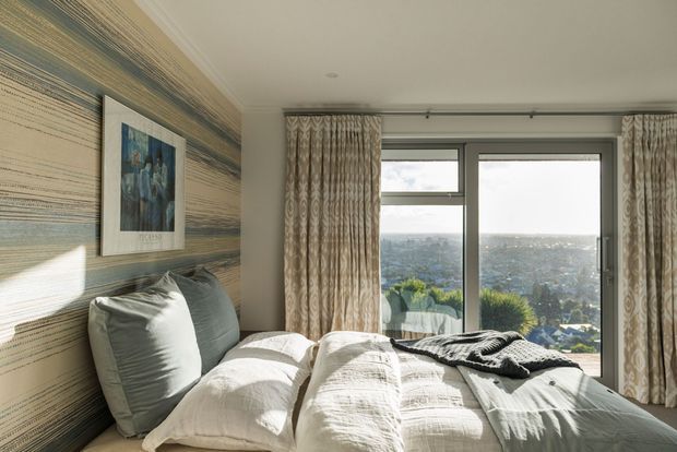 A bright, cozy bedroom with a bed positioned beside large floor-to-ceiling windows overlooking a distant landscape. The room features layered neutral bedding, oversized pillows, patterned full-length curtains, a textured horizontal-stripe feature wall, and framed artwork. Natural light creates a warm, relaxed atmosphere.