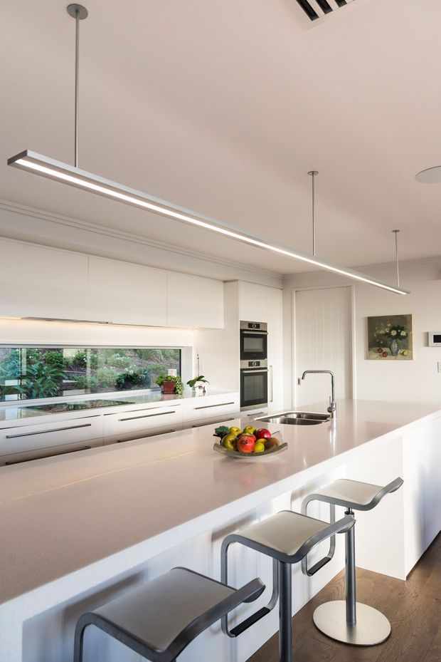 Bright contemporary white kitchen with extensive full-height cabinetry, a large pale island with integrated sink and raised breakfast bar, three modern bar stools, built-in oven, and linear pendant lighting. A long horizontal window provides views to greenery, while light timber flooring and minimal detailing create a clean, airy feel.