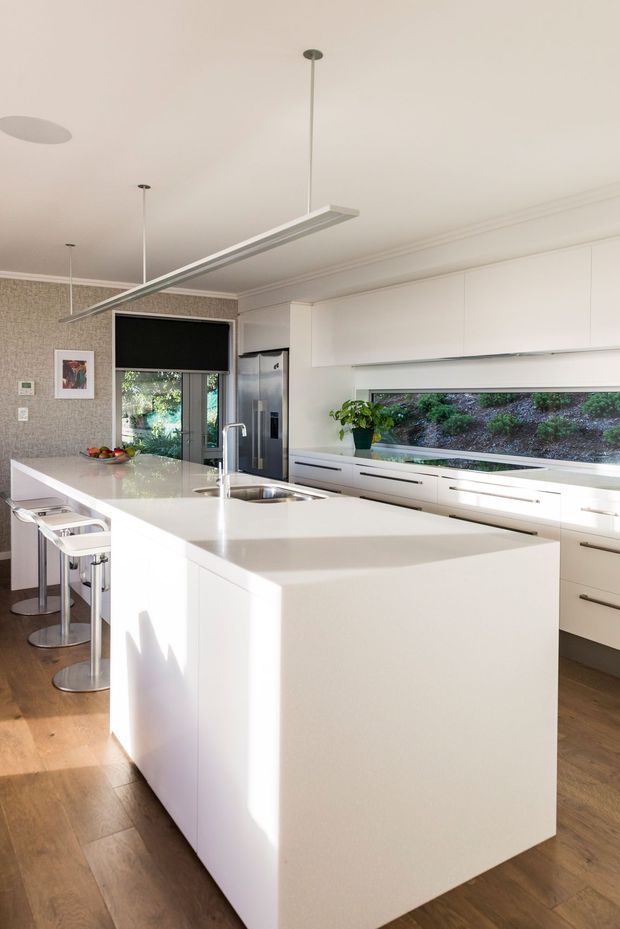 Bright contemporary kitchen with a large white waterfall island, integrated sink and seating for several people. The room has extensive white cabinetry, a long horizontal garden-view window, stainless-steel appliances, timber flooring, patterned feature wallpaper, and slim suspended linear lighting.