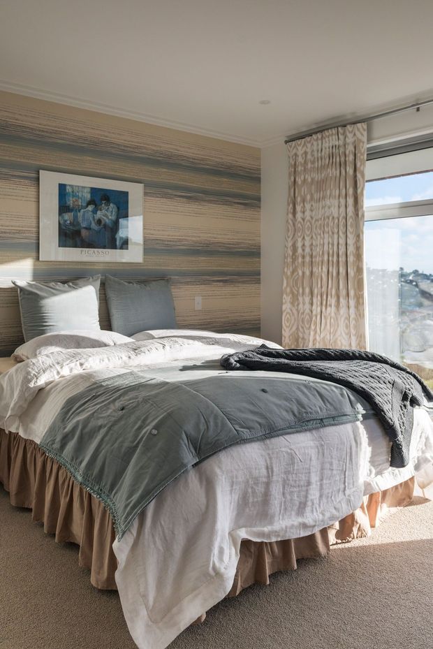 A bright, softly styled bedroom featuring a large upholstered bed layered with white and grey bedding, textured throw blankets, and multiple pillows. The bed is positioned against a horizontally patterned feature wall with framed artwork, while full-height patterned curtains frame a large window overlooking a distant urban view. Neutral tones, carpeted flooring, and abundant natural light create a calm, refined atmosphere.