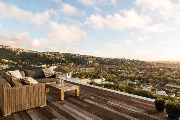 A spacious elevated outdoor deck or terrace overlooking a densely settled hilly suburban landscape. The deck features dark timber flooring, a woven outdoor sectional sofa with patterned cushions, a rustic timber coffee table, glass balustrading, and potted plants. Warm late-afternoon light creates a relaxed, scenic entertaining area.