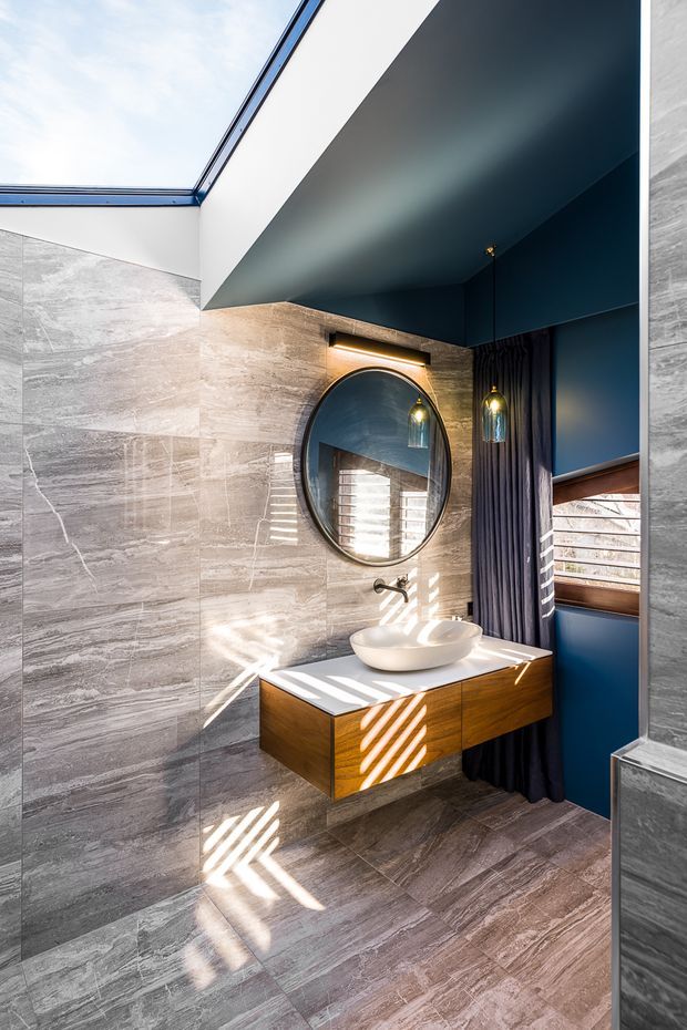 A contemporary bathroom with a floating timber vanity, white vessel basin, round framed mirror, wall-mounted tapware, and dark blue ceiling and wall accents. Large grey stone-look tiles cover the walls and floor. A skylight and timber-framed horizontal window bring in strong natural light, creating striped shadows across the vanity and floor. Dark curtains and two pendant lights add contrast.