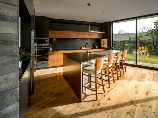 A contemporary open-plan kitchen with a large dark island and integrated seating for five, set against warm timber cabinetry and a dark backsplash. The room features wide-plank timber flooring, full-height sliding glass doors opening to a garden, minimalist pendant lighting, integrated appliances, and a concrete-toned wall. Strong natural light creates a warm, refined atmosphere.