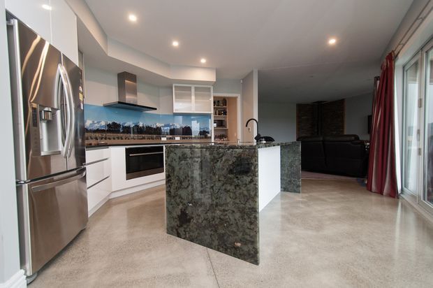 Open-plan contemporary kitchen and living area with white cabinetry, stainless-steel appliances, a large dark veined stone island, integrated cooktop and rangehood, and polished concrete flooring. Floor-to-ceiling glazing and red curtains open toward the living zone with a dark sofa.