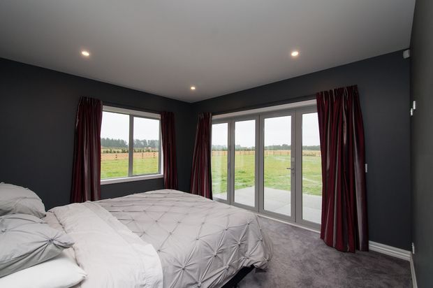 A dark, contemporary bedroom with charcoal walls, a low white ceiling with recessed downlights, grey carpet, and a large bed dressed in white and pale grey bedding. Deep burgundy curtains frame a side window and wide glazed French doors opening to a rural grassy outlook.