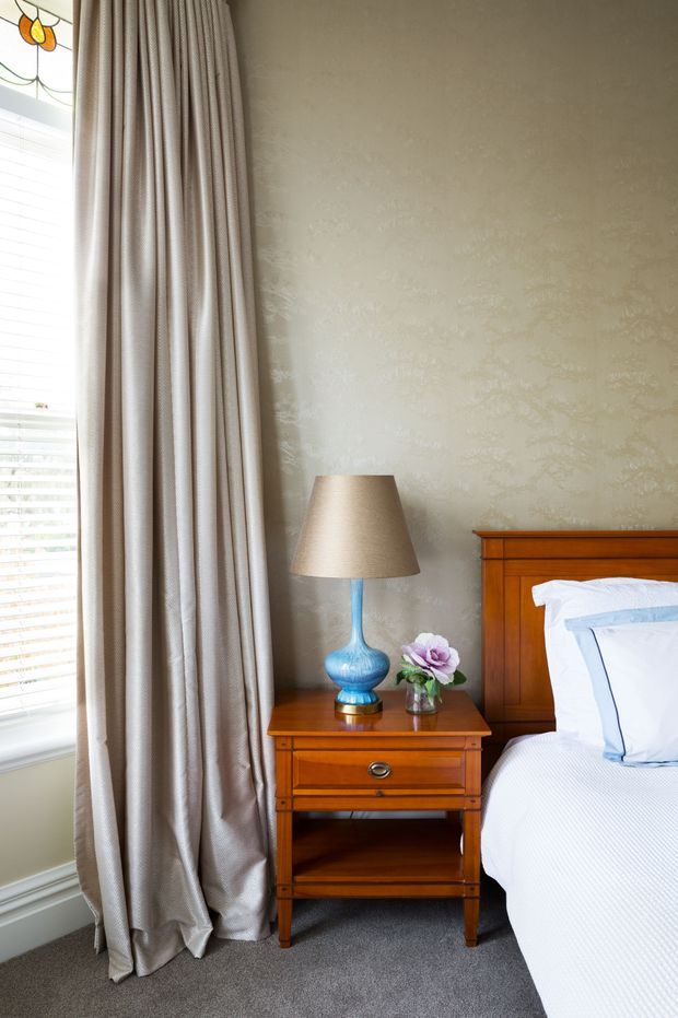 A traditional bedroom corner featuring a wooden bed headboard and matching bedside table, a blue ceramic lamp, pale patterned wallpaper, full-length taupe curtains, a bright window with blinds, and grey carpet. The styling is refined and classic with warm timber furniture and soft neutral textiles.
