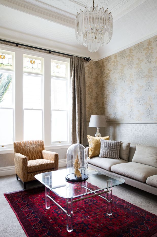 Elegant traditional living room with ornate decorative plaster ceiling cornices, patterned floral wallpaper, stained-glass upper window details, and a large crystal chandelier. A neutral upholstered sofa and patterned armchair surround a clear glass coffee table with chrome supports, set over a richly coloured red Persian-style rug. The room has a refined, layered, classic character with abundant natural light.