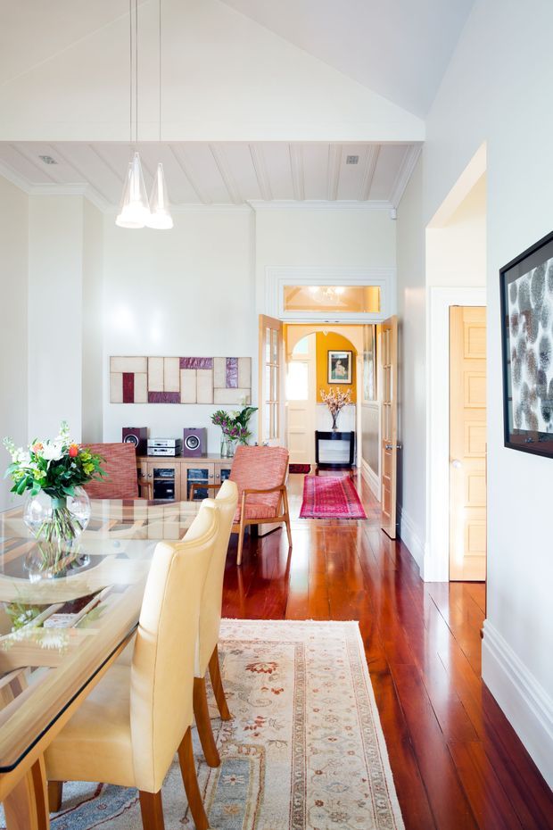 Bright, elegant open-plan dining and living area with polished dark timber flooring, a glass dining table surrounded by upholstered cream chairs, and a compact timber media console with stereo equipment. The room features pale walls, detailed white coffered and panelled ceiling elements, a high pitched ceiling, pendant lights, framed artwork, and a view through glazed double doors into a warm-toned hallway or adjoining room. Layered traditional rugs, floral arrangements, and warm wood accents create a refined, classic interior.